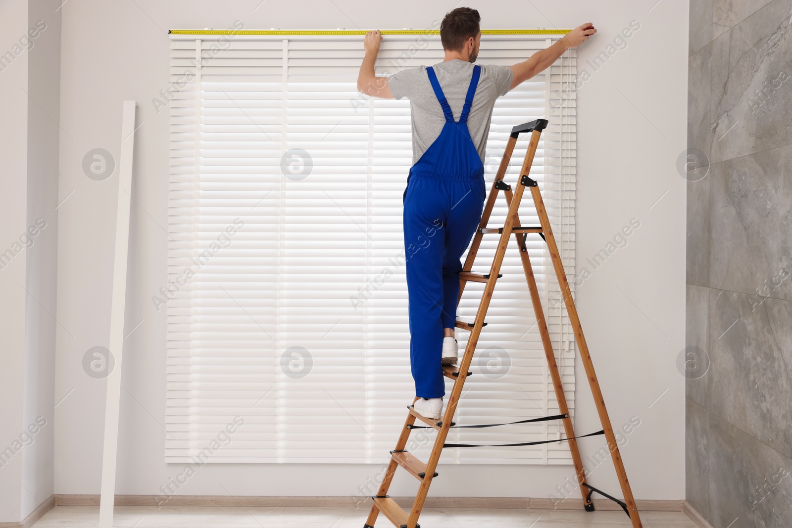 Photo of Worker in uniform using measuring tape while installing horizontal window blinds on stepladder indoors, back view