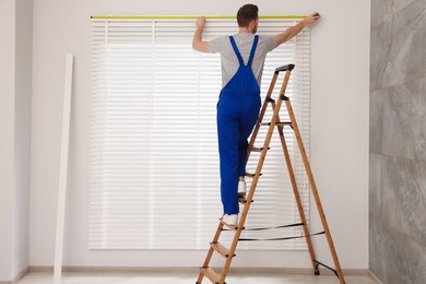 Worker in uniform using measuring tape while installing horizontal window blinds on stepladder indoors, back view