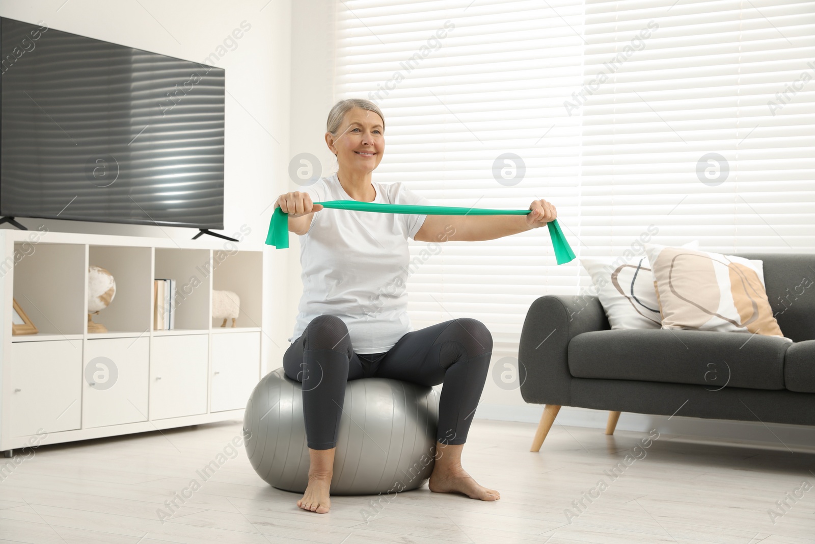 Photo of Senior woman doing exercise with elastic resistance band on fitness ball at home