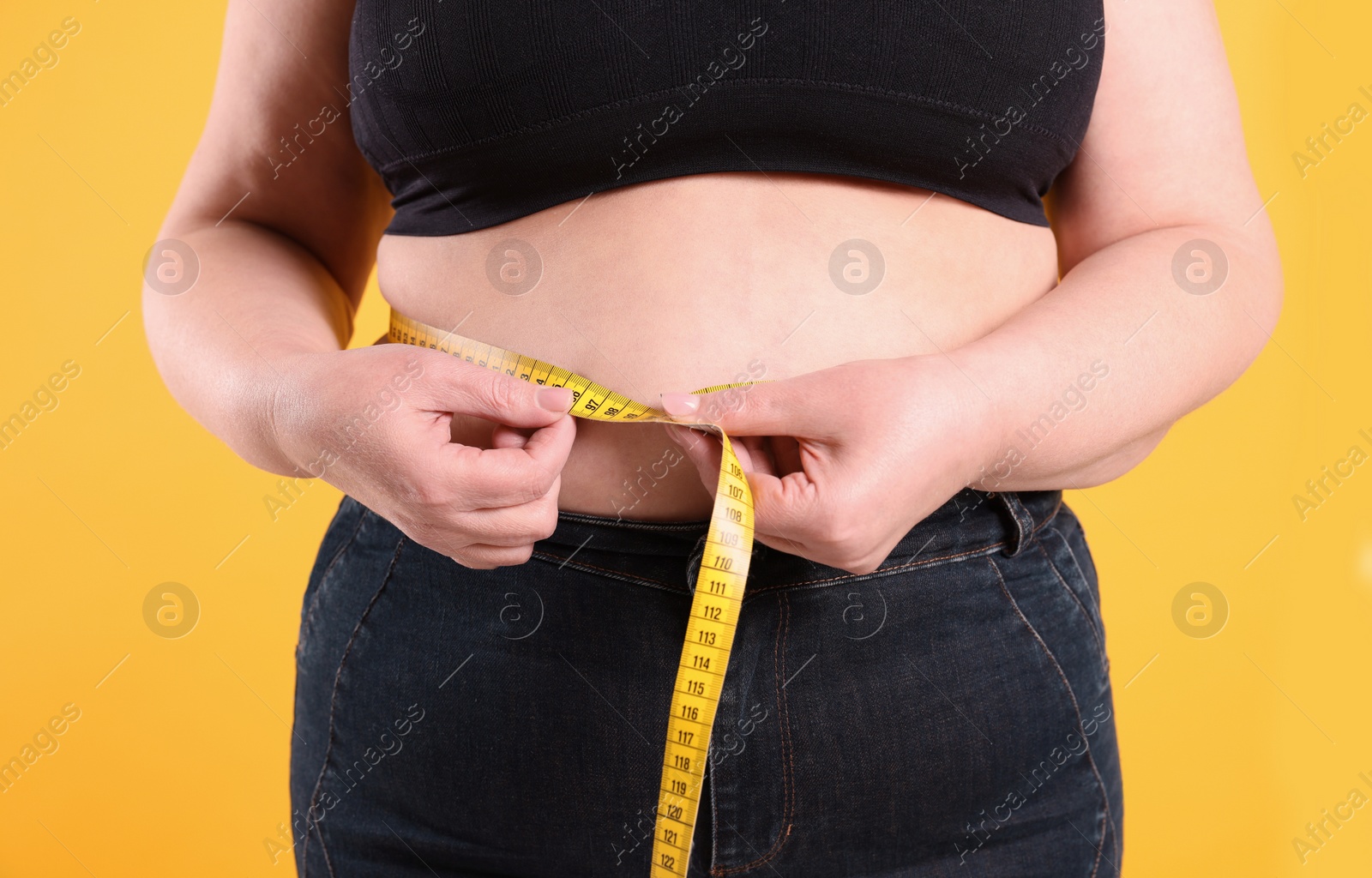 Photo of Overweight woman measuring waist with tape on yellow background, closeup