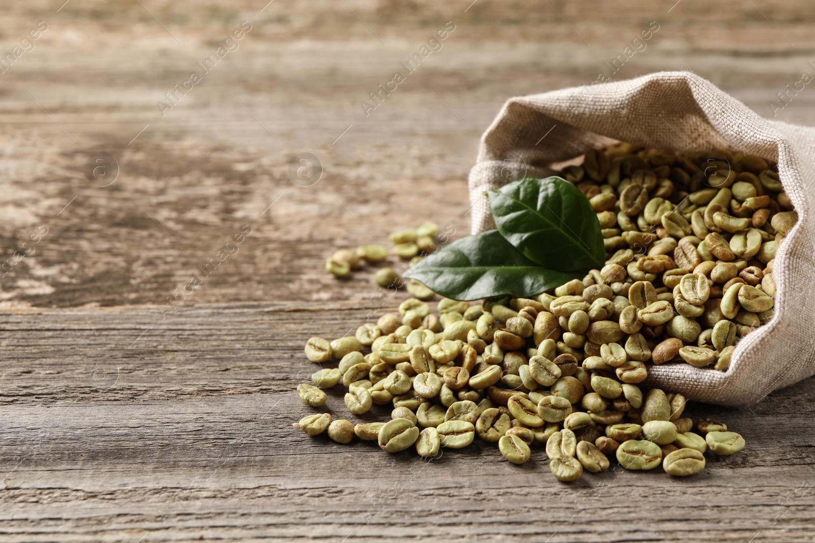 Photo of Overturned sackcloth bag with green coffee beans and leaves on wooden table, closeup. Space for text