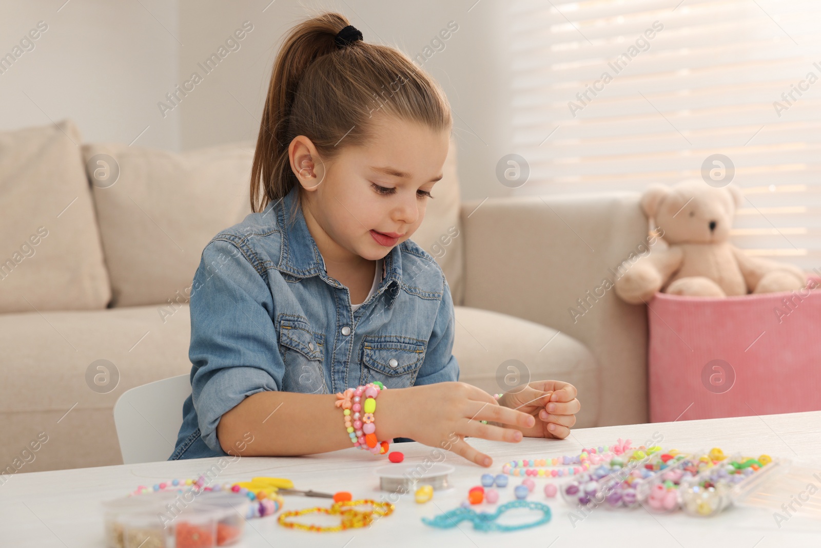 Photo of Cute little girl making beaded jewelry at table in room