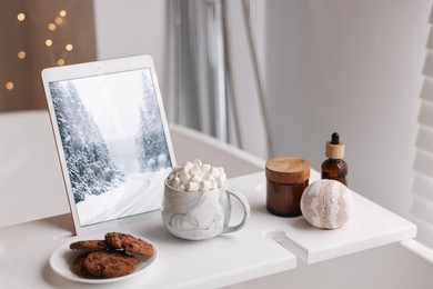 Photo of White wooden tray with tablet, cookies and spa products on bathtub in bathroom
