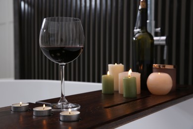 Photo of Glass of tasty red wine and burning candles on bathtub tray