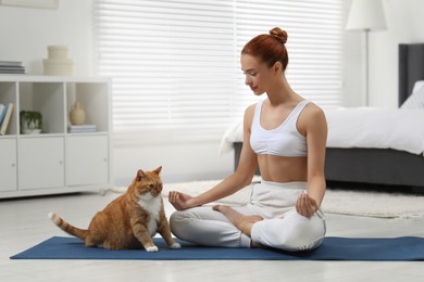 Beautiful woman with cute red cat practicing yoga on mat at home