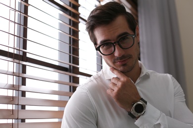 Photo of Handsome young man in white shirt with glasses standing near window indoors
