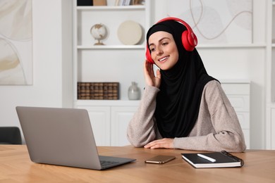 Muslim woman in hijab using laptop at wooden table in room