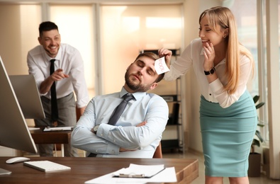 Photo of Young woman sticking note with word Fool to colleague's face in office. Funny joke