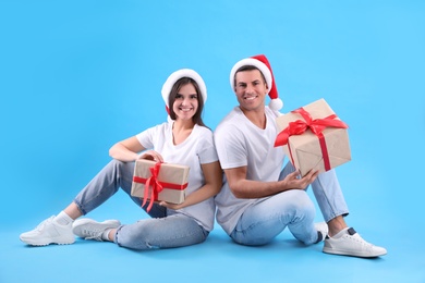 Photo of Beautiful happy couple in Santa hats sitting with Christmas gifts on light blue background