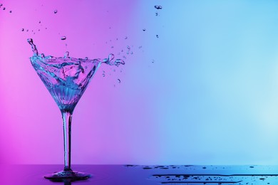 Glass of martini and splashes on table in neon lights, space for text