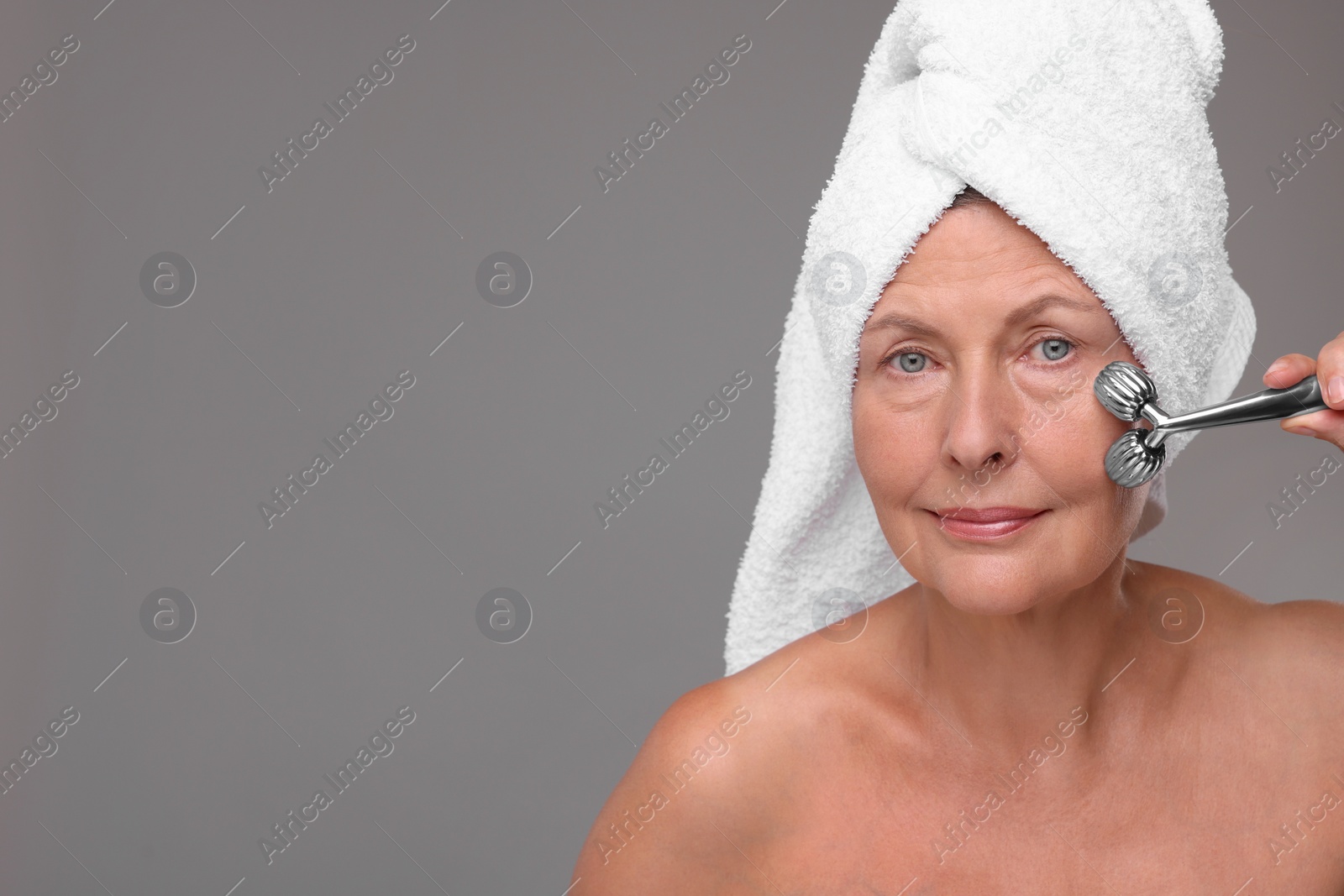 Photo of Woman massaging her face with metal roller on grey background, space for text