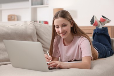 Photo of Online learning. Teenage girl writing summary near laptop on sofa at home