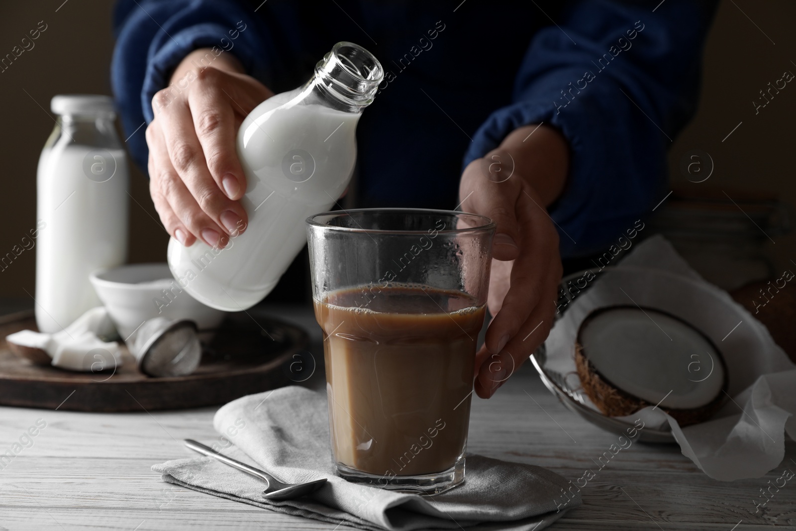 Photo of Woman holding bottle with coconut milk near glass of coffee at white wooden table, closeup