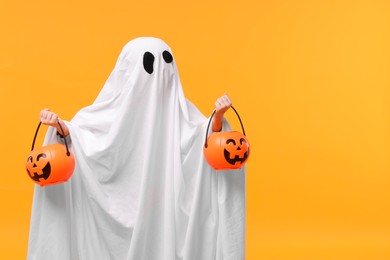 Child in white ghost costume holding pumpkin buckets on orange background, space for text. Halloween celebration