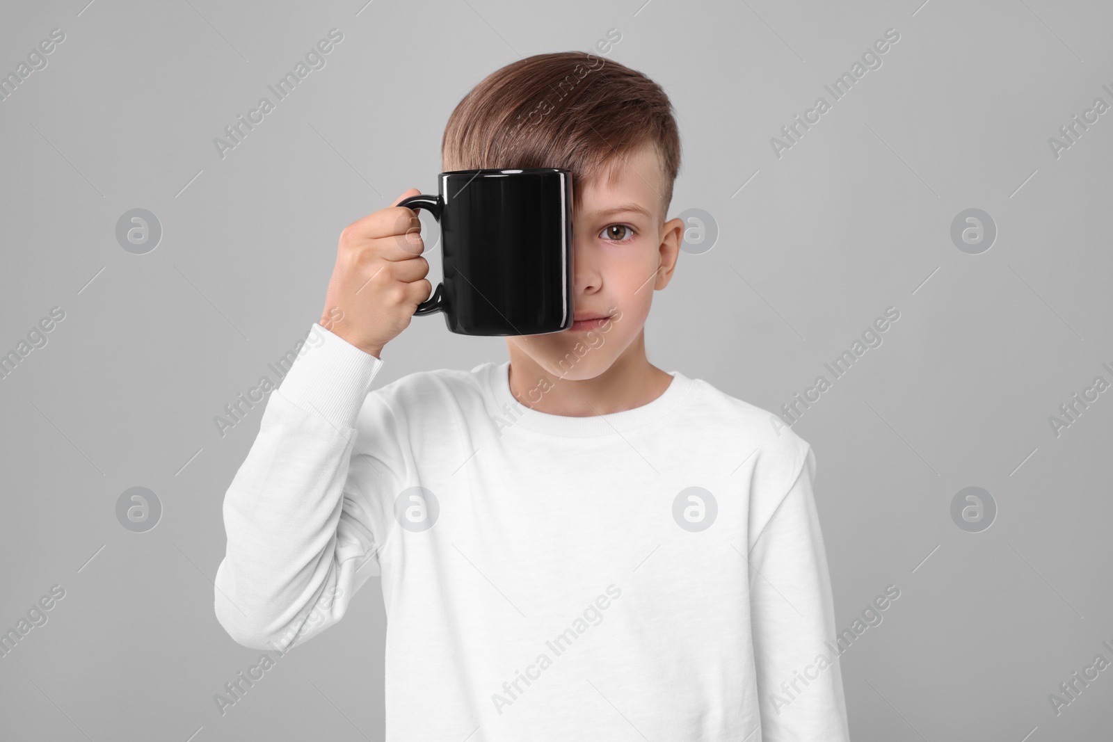 Photo of Cute boy covering eye with black ceramic mug on light grey background