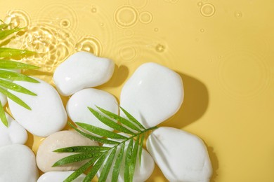 Spa stones and palm leaves in water on yellow background, flat lay