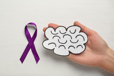 Woman holding paper brain cutout near purple ribbon on white wooden background, top view. Epilepsy awareness