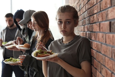 Photo of Young poor woman and other people with food at brick wall indoors