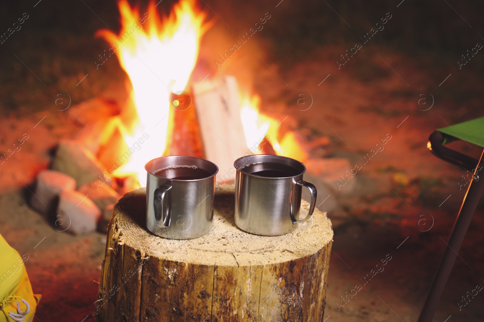 Photo of Cups of hot drink on old log near bonfire outdoors. Camping season
