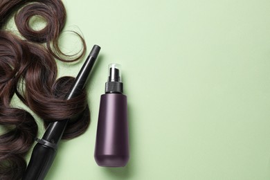 Photo of Spray bottle with thermal protection, lock of brown hair and stylish curling iron on pale green background, flat lay. Space for text