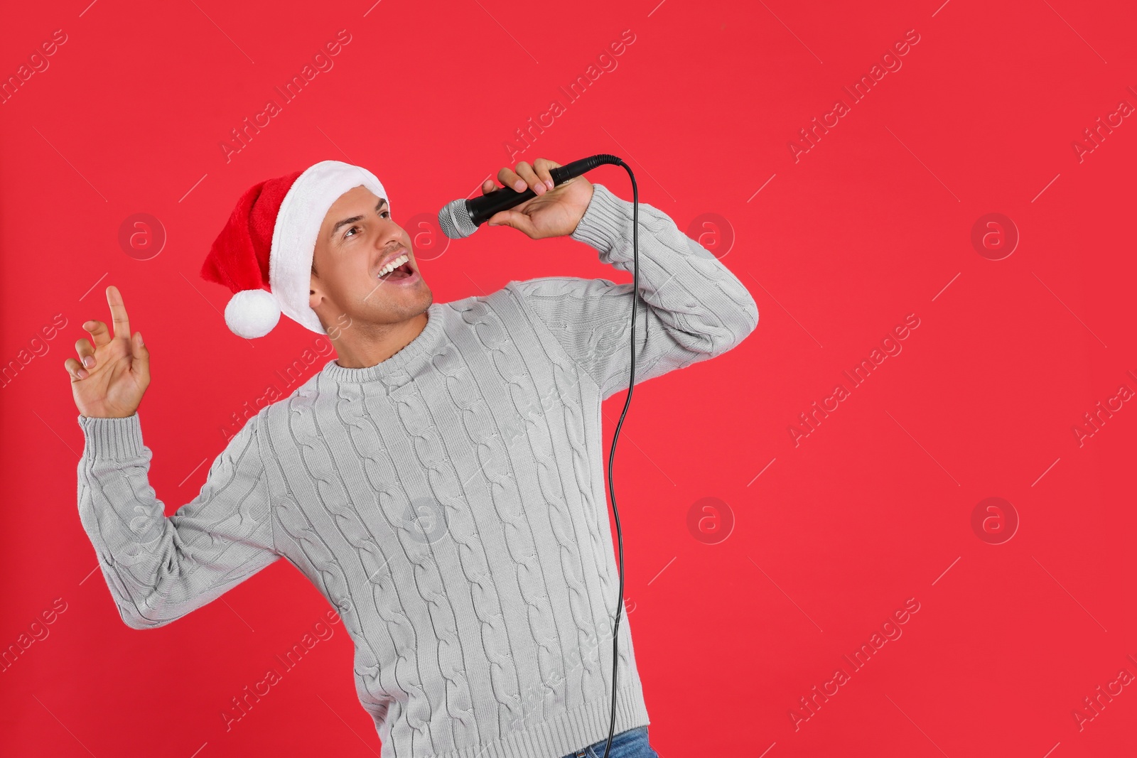 Photo of Happy man in Santa Claus hat singing with microphone on red background, space for text. Christmas music
