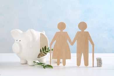 Photo of Pension savings. Figure of senior couple, stacked coins, piggy bank and green twigs on white wooden table