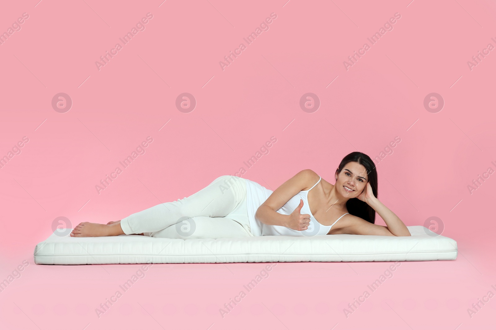 Photo of Young woman lying on soft mattress and showing thumbs up against pink background