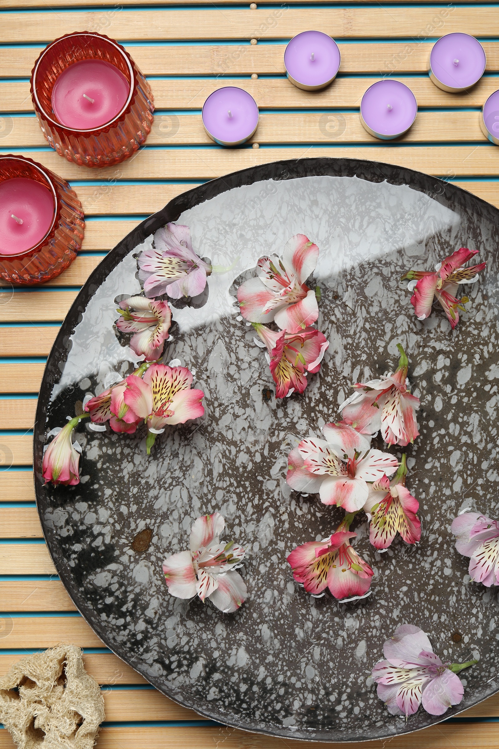 Photo of Bowl of water with flowers, candles and loofah on wooden mat, flat lay. Spa treatment