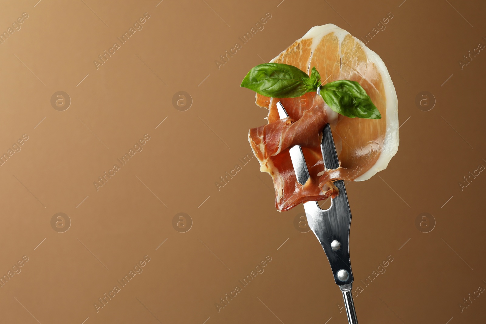 Photo of Fork with slice of tasty jamon and basil leaves on brown background, closeup. Space for text