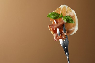 Fork with slice of tasty jamon and basil leaves on brown background, closeup. Space for text