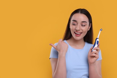 Photo of Happy young woman holding electric toothbrush and pointing on yellow background, space for text