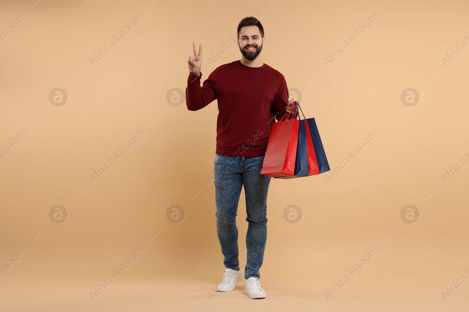 Photo of Smiling man with many paper shopping bags showing peace sign on beige background