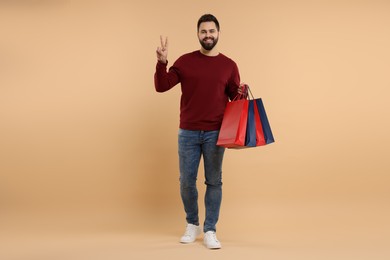 Photo of Smiling man with many paper shopping bags showing peace sign on beige background