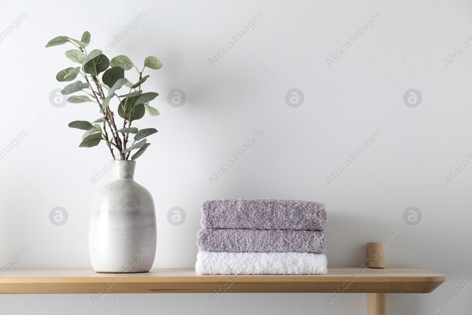 Photo of Stacked terry towels and eucalyptus branches in vase on wooden shelf near white wall
