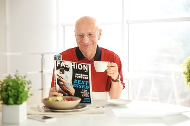 Photo of Elderly man reading magazine while having breakfast at home