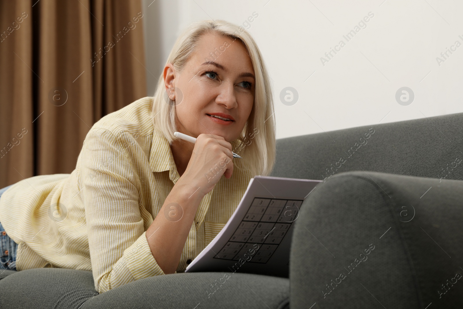 Photo of Middle aged woman solving sudoku puzzle on sofa at home