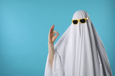 Stylish ghost. Woman covered with white sheet in sunglasses showing OK gesture on light blue background, space for text