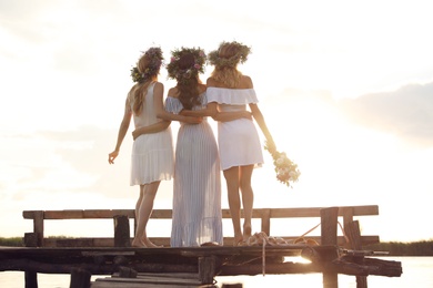 Photo of Young women wearing wreaths made of beautiful flowers on pier near river, back view