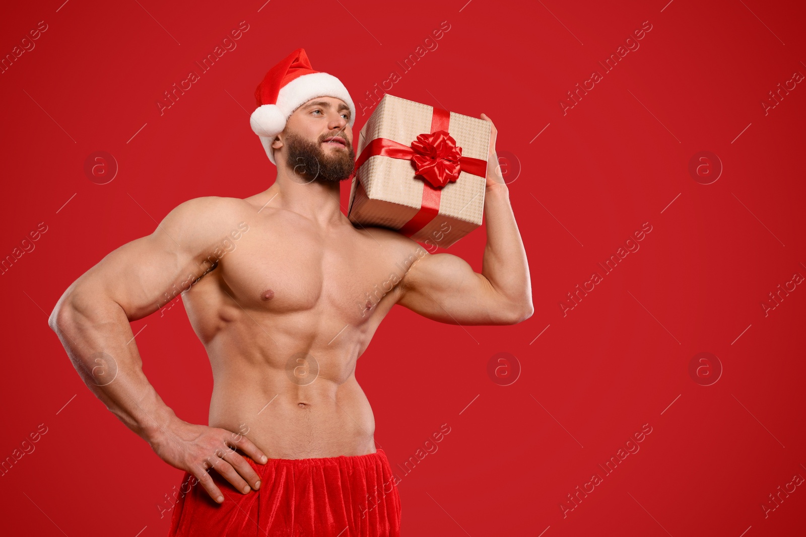 Photo of Attractive young man with muscular body in Santa hat holding Christmas gift box on red background, low angle view. Space for text