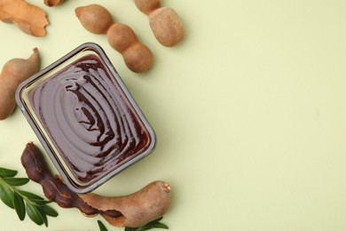 Photo of Bowl of tasty tamarind sauce, leaves and fresh pods on light green table, flat lay. Space for text
