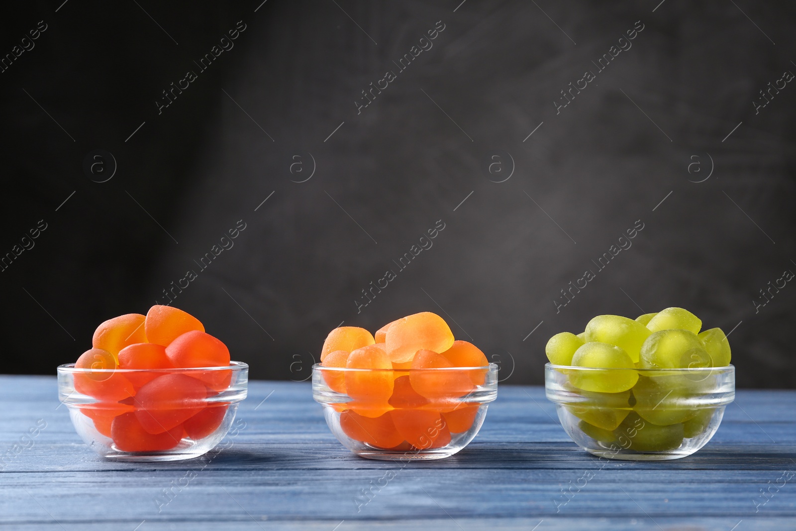 Photo of Glass bowls with tasty jelly candies on blue wooden table against grey background, space for text