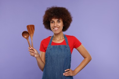 Photo of Happy young woman in apron holding spoon and spatula on purple background