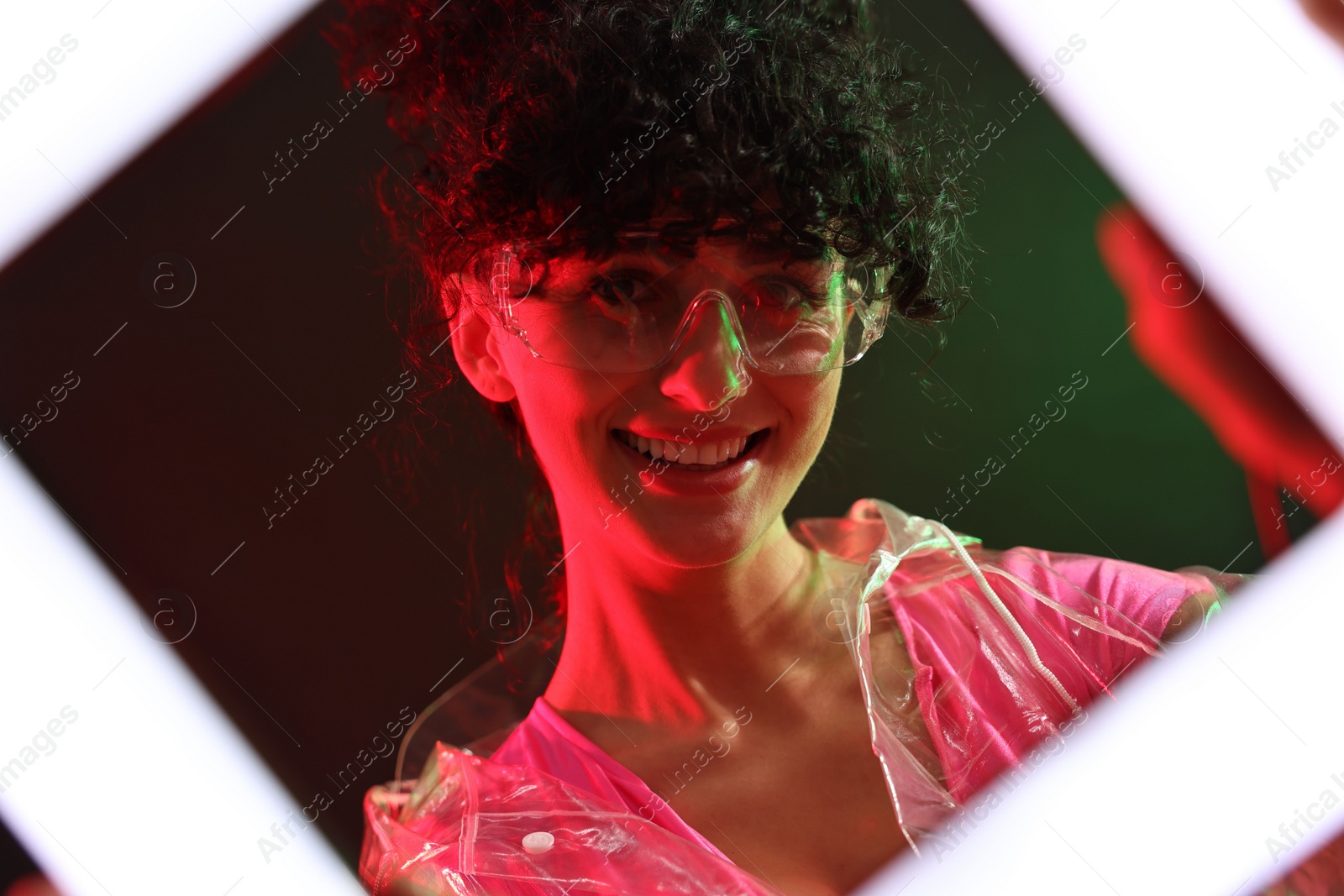 Photo of Beautiful young woman in transparent coat and sunglasses with square lamp on color background in neon lights