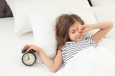 Photo of Cute little girl with alarm clock awaking in bed. Healthy sleep