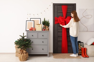 Photo of Woman decorating wooden door with red bow indoors. Christmas mood