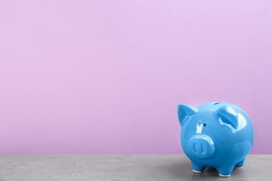 Photo of Blue piggy bank on light grey table against violet background. Space for text