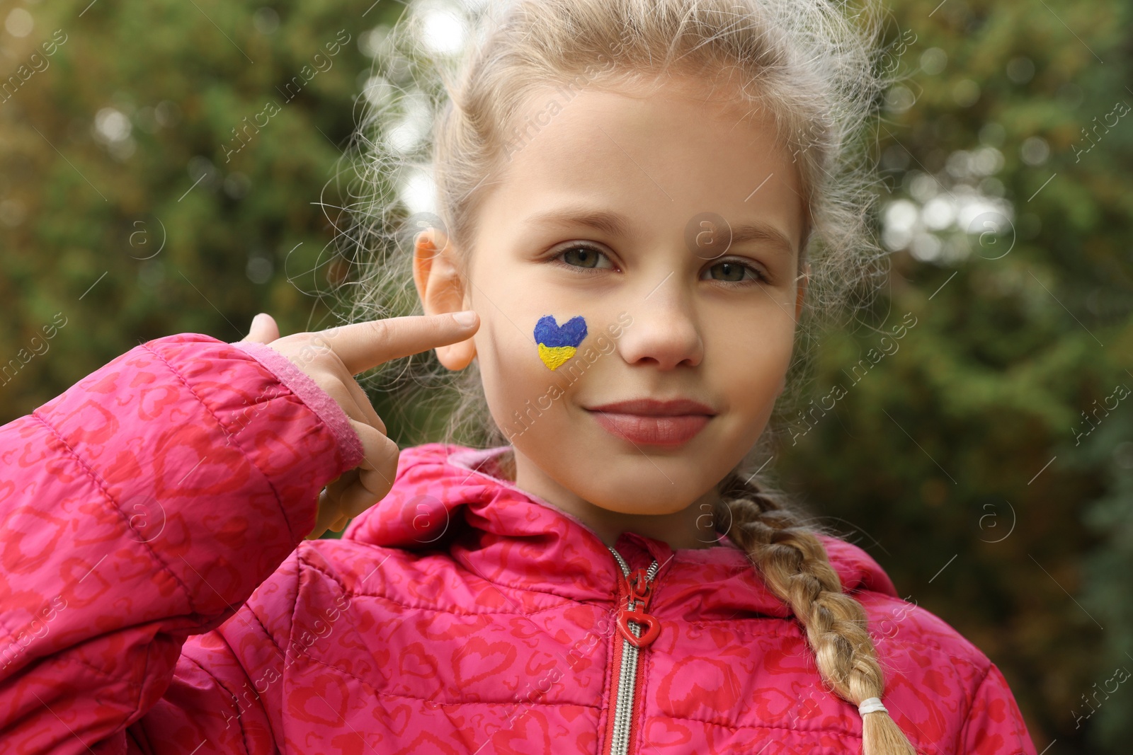 Photo of Little girl with drawing of Ukrainian flag on face in heart shape outdoors