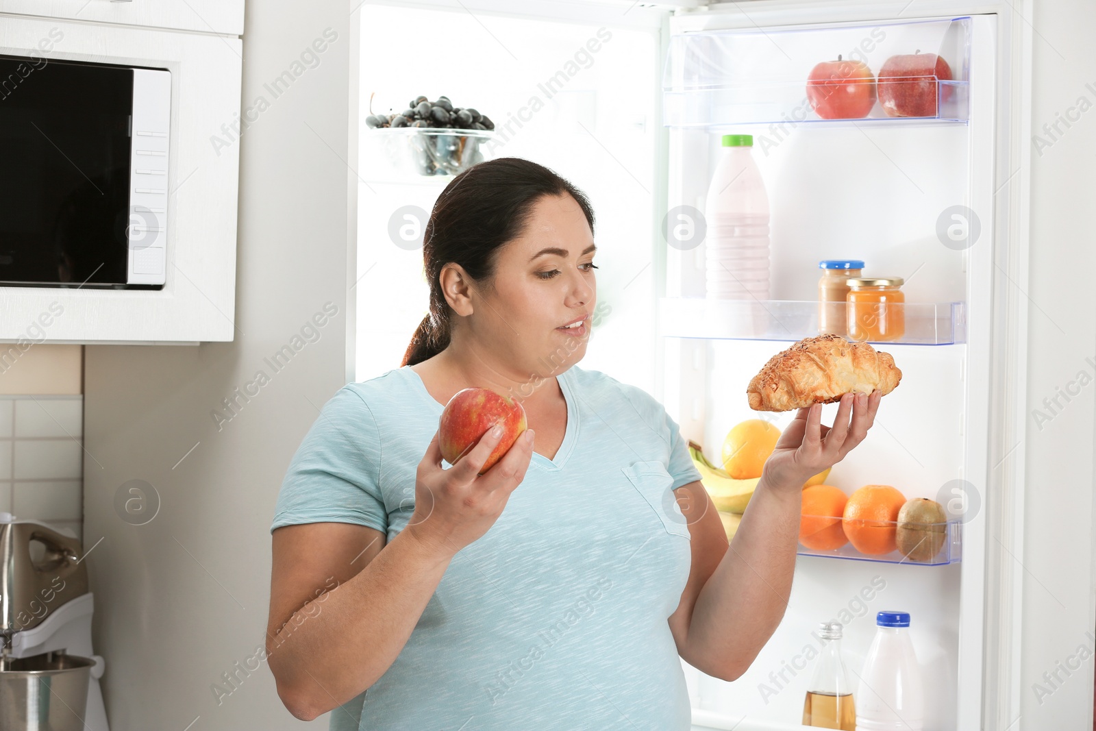 Photo of Woman choosing between apple and croissant near fridge at kitchen. Healthy diet