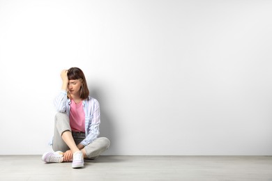 Photo of Unhappy young girl sitting on floor near white wall. Space for text