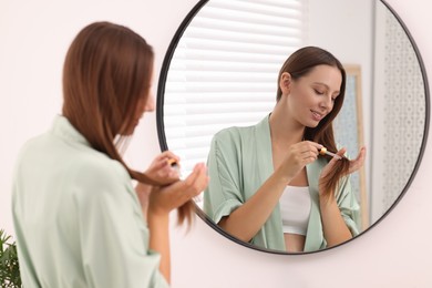 Photo of Beautiful woman applying serum onto hair near mirror indoors